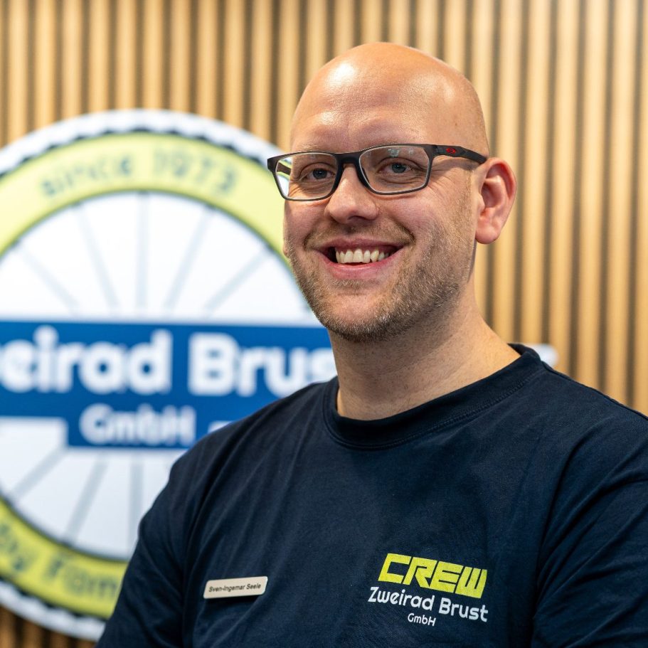 Lächelnder Mann mit Brille, trägt ein T-Shirt mit dem Aufdruck „Crew“. Hintergrund mit Logo.